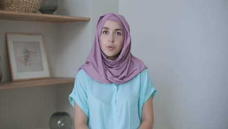 Una-Mujer-Musulmana-Con-Hijab-Se-Sienta-En-Una-Mesa-Y-Mira-A-La-Cámara-Agitando-La-Mano-Y-Hablando.-Vlog-O-Videollamada-A-Una-Mujer-Con-Hijab.-Foto-De-La-Cabeza-De-Una-Mujer-árabe-En-Conversación.